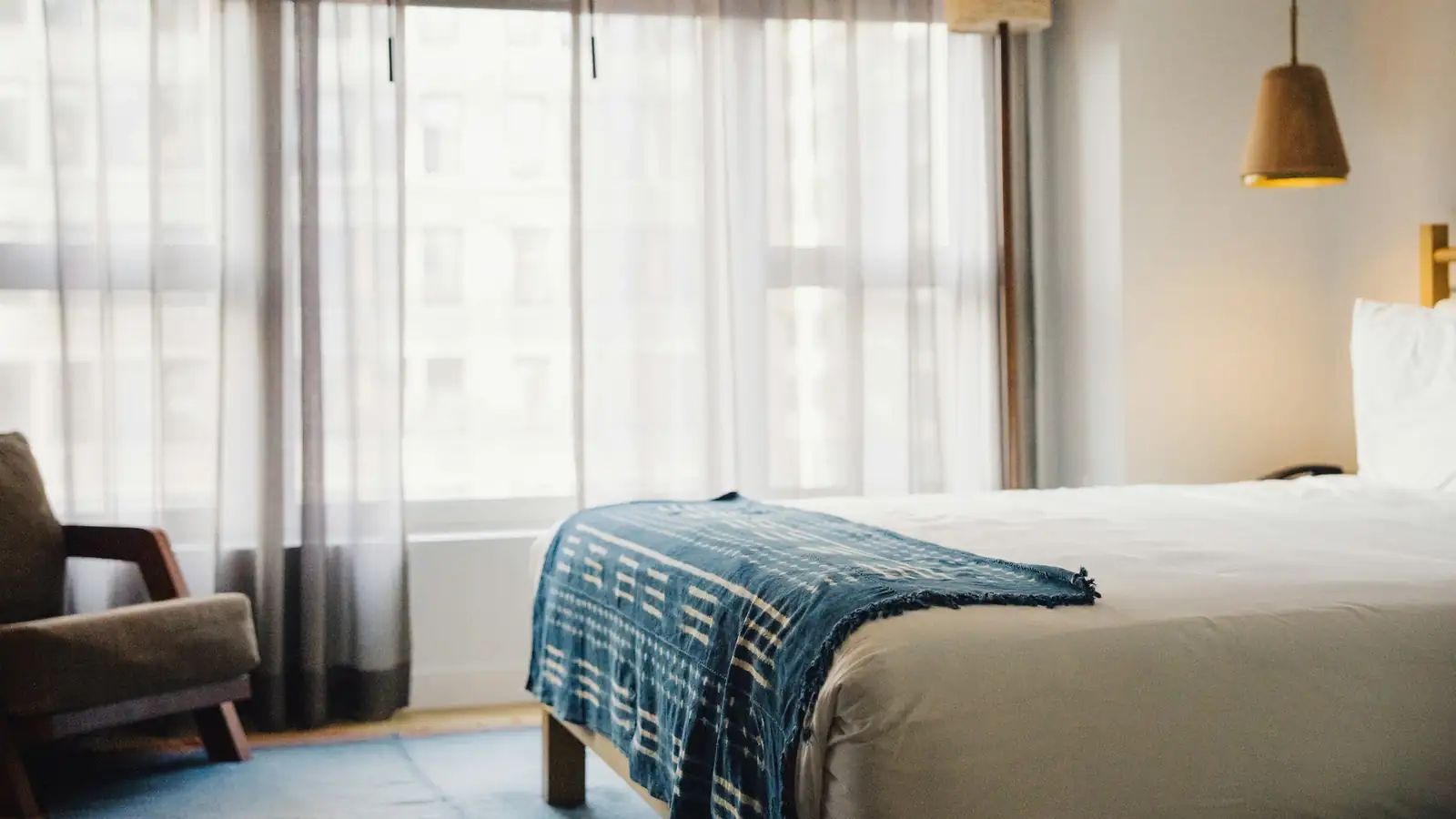 Quarto de hotel iluminado pela janela, com cama arrumada, manta azul e poltrona