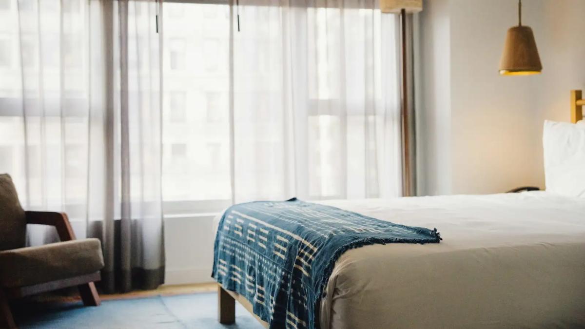 Quarto de hotel iluminado pela janela, com cama arrumada, manta azul e poltrona