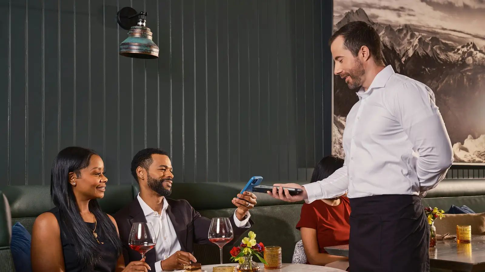 Garçom recebendo o pagamento pelo celular de um casal à mesa do restaurante
